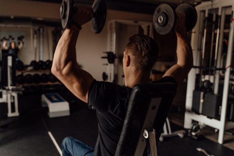 Ein muskulöser Mann, von hinten gesehen, der ein schwarzes T-Shirt trägt, auf einer verstellbaren Bank sitzt und in einem Fitnessstudio zwei schwere Hanteln über den Kopf drückt