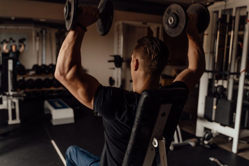 Ein muskulöser Mann, von hinten gesehen, der ein schwarzes T-Shirt trägt, auf einer verstellbaren Bank sitzt und in einem Fitnessstudio zwei schwere Hanteln über den Kopf drückt